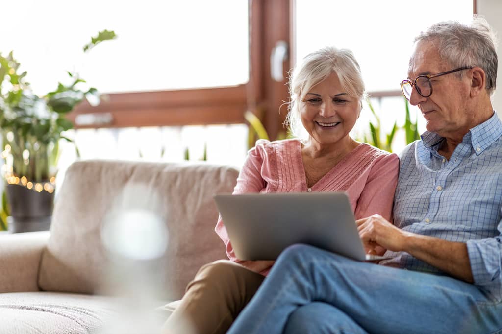 Older couple sit on a sofa browsing a laptop together in a bright, plant-filled room