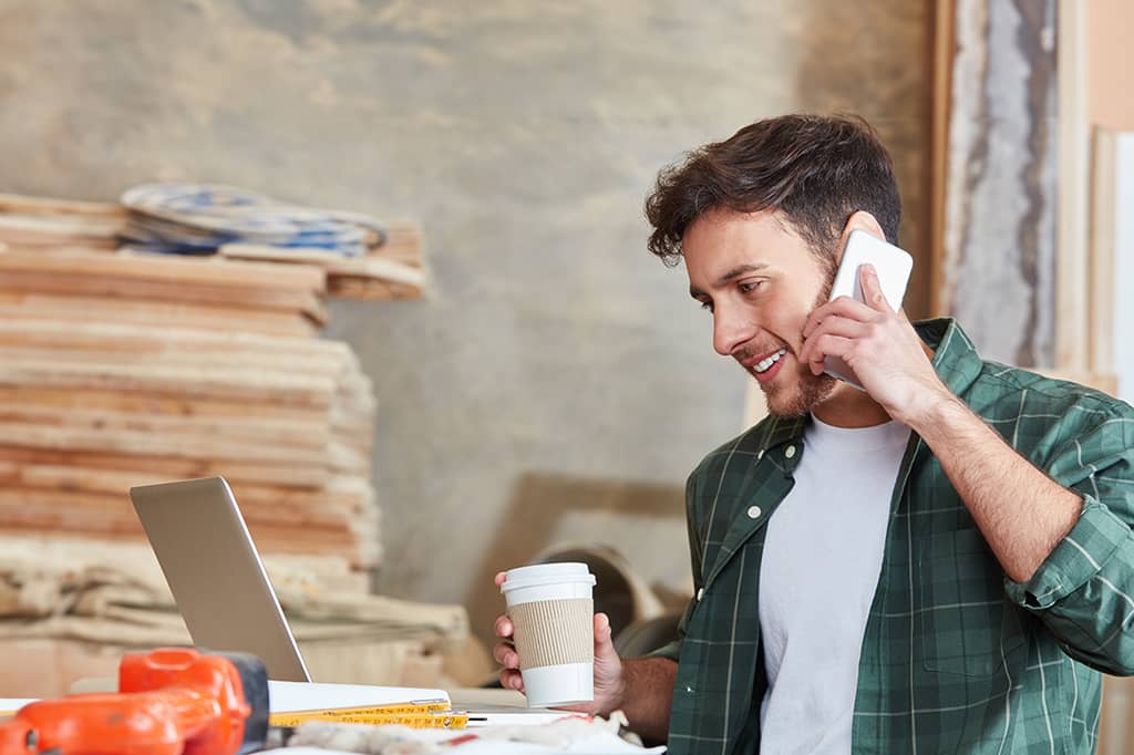 Self-employed tradesman takes a phone call at his workshop laptop with a coffee