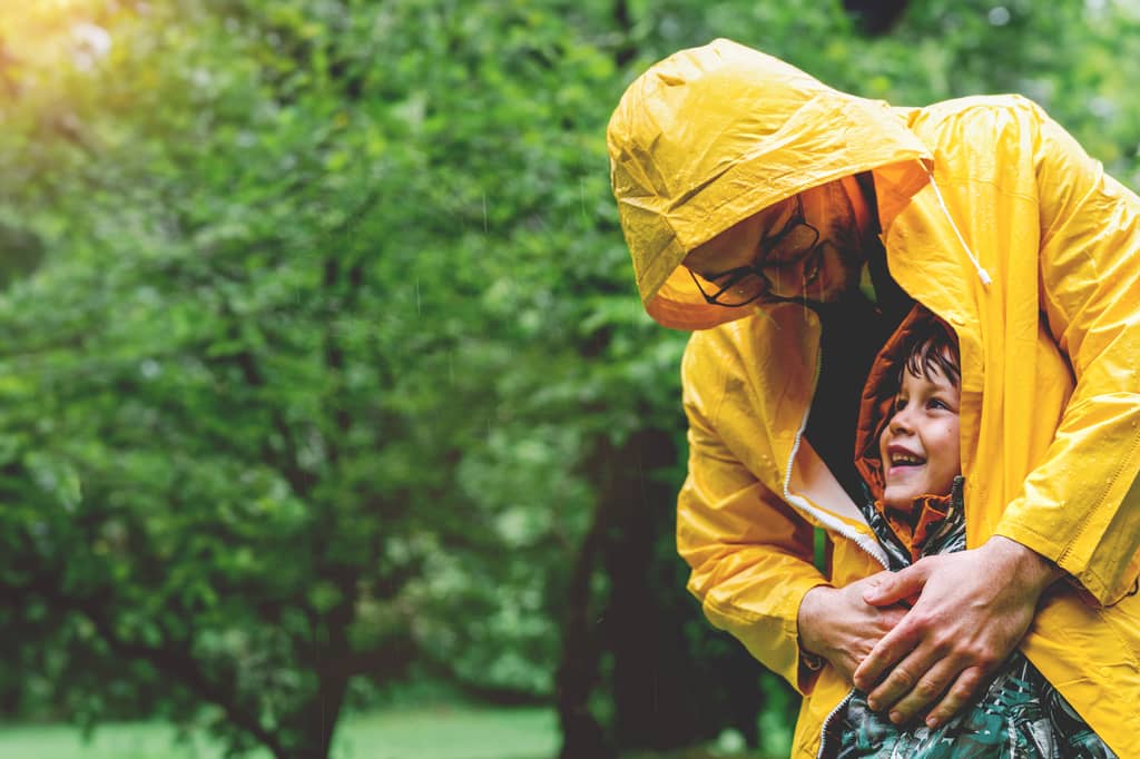 Parent in a yellow raincoat shelters a smiling child from the rain among green trees