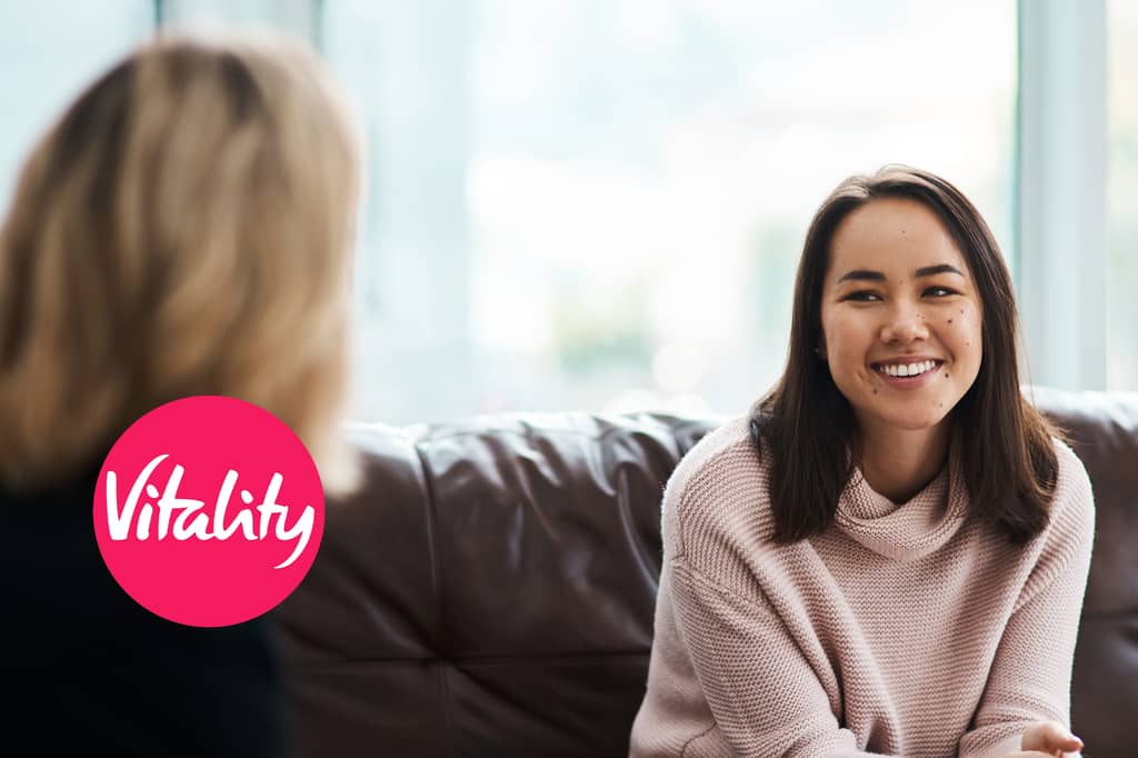 Smiling woman talks with another person on a sofa, with the Vitality logo overlaid