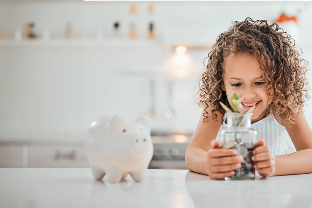 Young girl smiling at a jar of coins and a plant beside a piggy bank