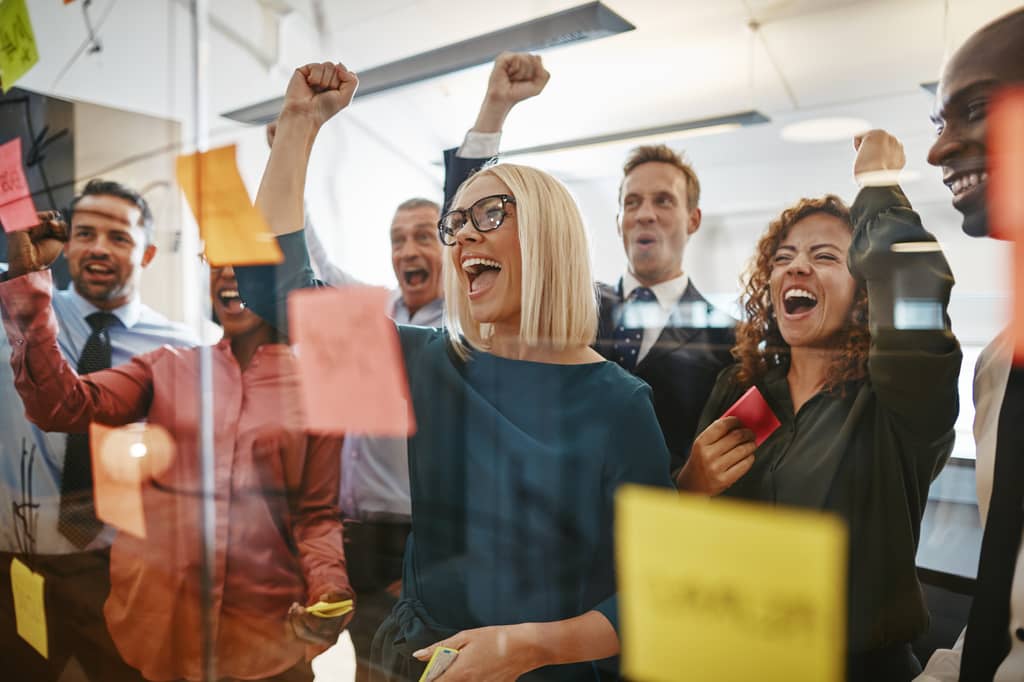 Office team cheer and raise their fists behind a glass wall of sticky notes