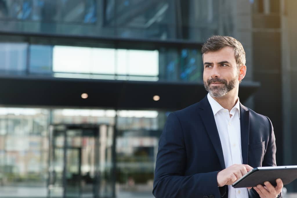 Company director in a dark suit using a tablet outside a glass office building
