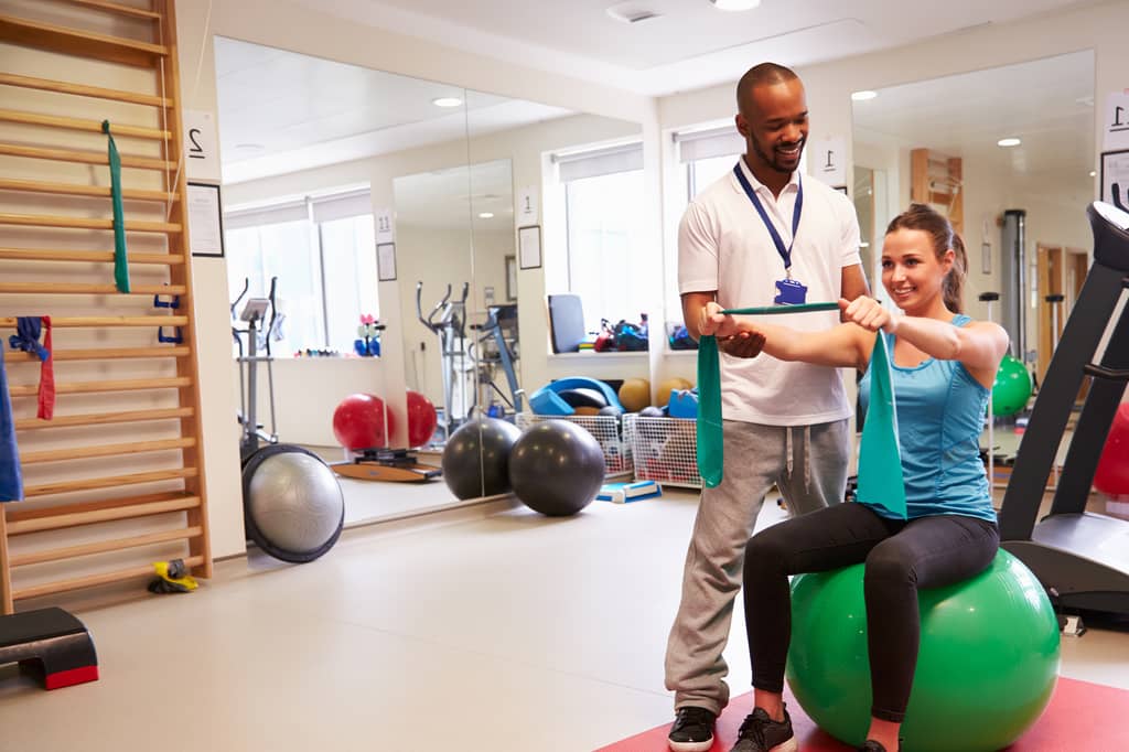 Physiotherapist guides a woman doing resistance band exercises on a gym ball during rehab