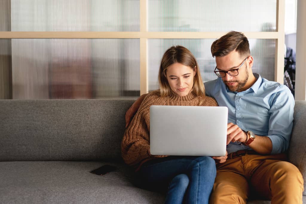 Young couple sit on a sofa looking at a laptop, working through health insurance details