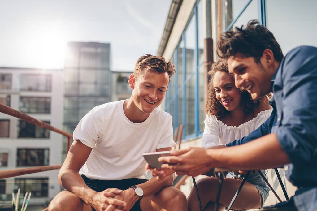 Three young friends laughing together on a rooftop terrace while looking at a phone