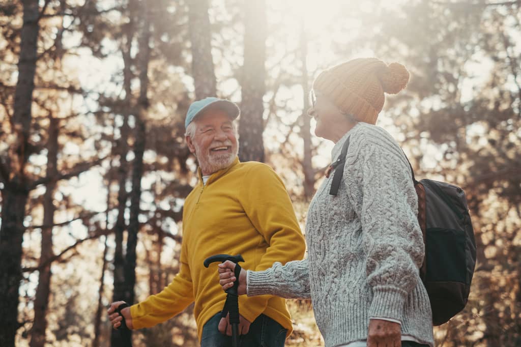 Older couple laugh together while hiking with walking poles through sunlit woodland