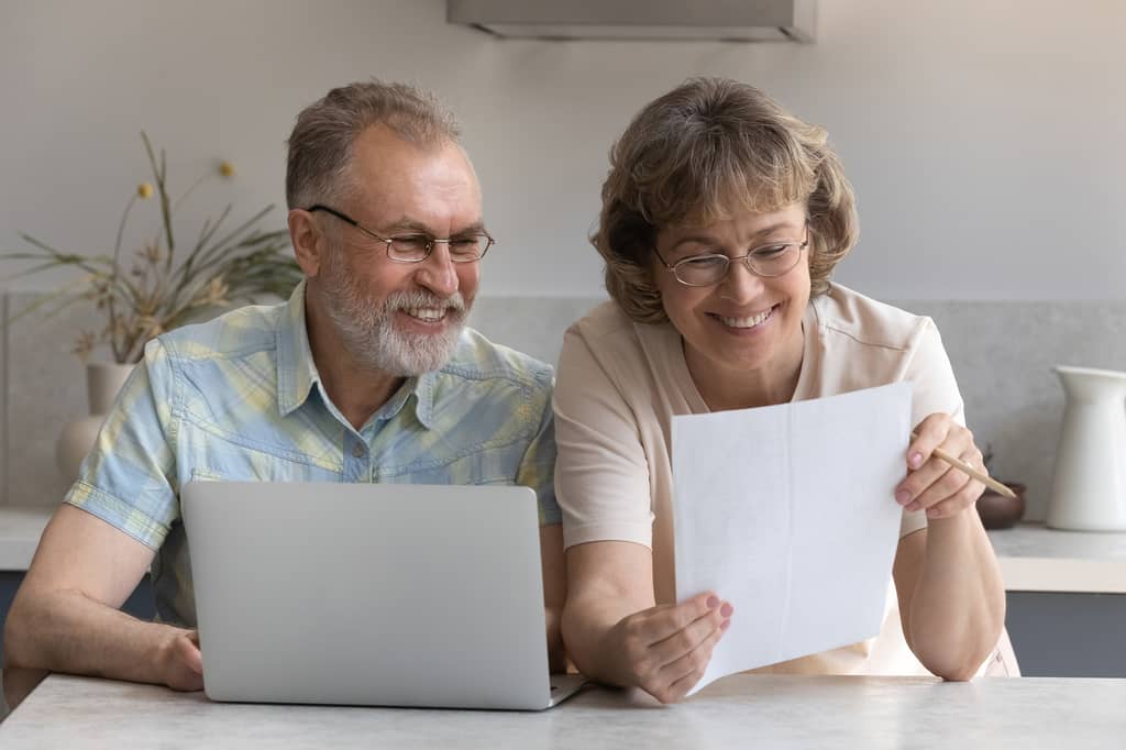 Older couple smile while reviewing a document and laptop at the kitchen counter