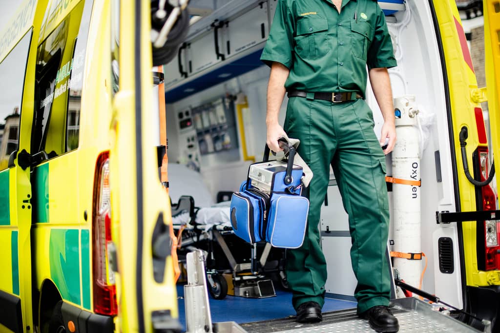 Paramedic in green uniform stands in an ambulance holding a medical bag beside an oxygen cylinder