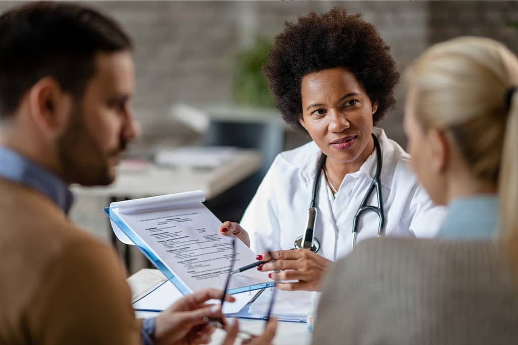 Doctor with a stethoscope discussing a policy document with a couple in a consultation