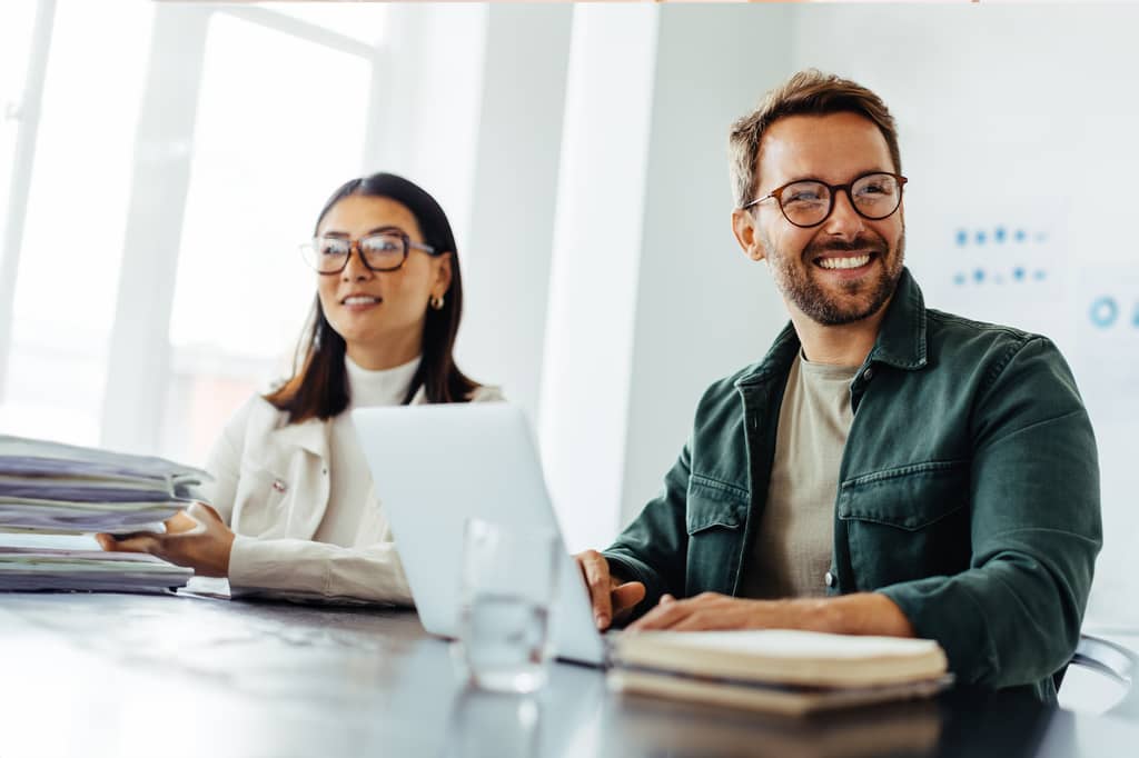 Two colleagues smiling at a desk with a laptop and folders in a bright office