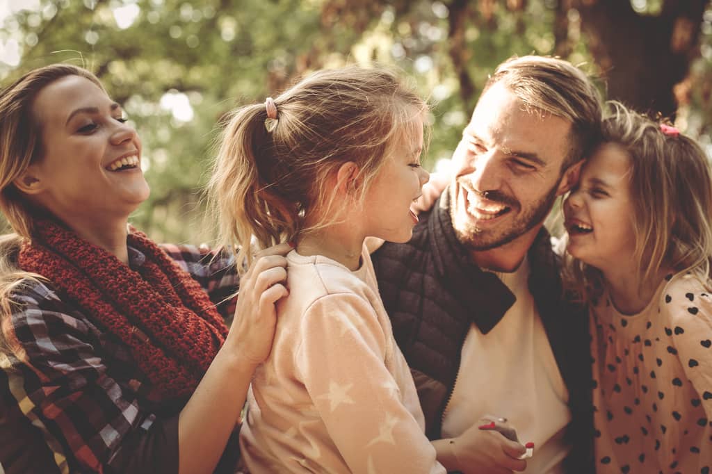 Parents and two young daughters laugh together outdoors among autumn trees