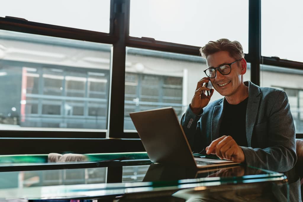 Businessman in a suit smiling on the phone while using a laptop at a glass desk