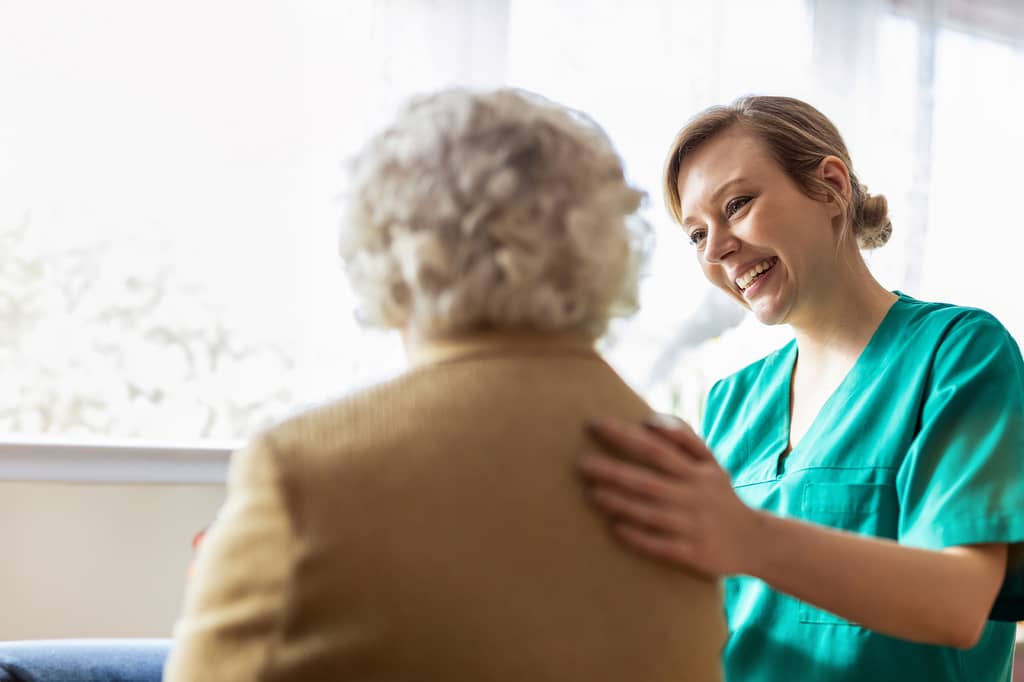 Nurse in green scrubs smiles and rests a hand on an elderly patient's shoulder