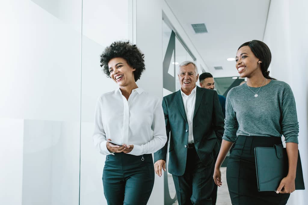Three smiling business colleagues walk and talk together along an office corridor