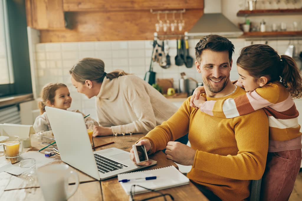 Father and daughter at a kitchen table with a laptop while mum and child chat behind