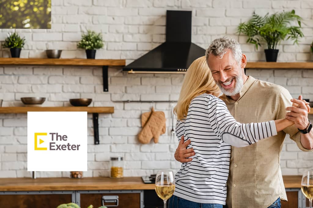 Couple dance and laugh in their kitchen, with The Exeter logo overlaid