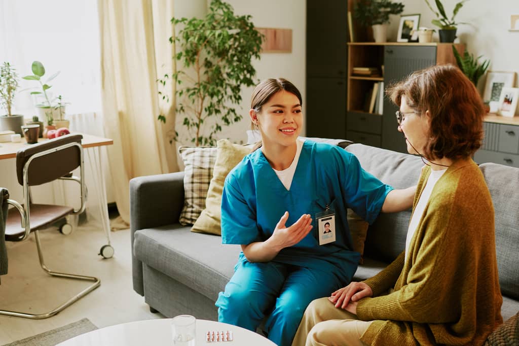 Nurse in blue scrubs talks with an older woman on a sofa during a home consultation