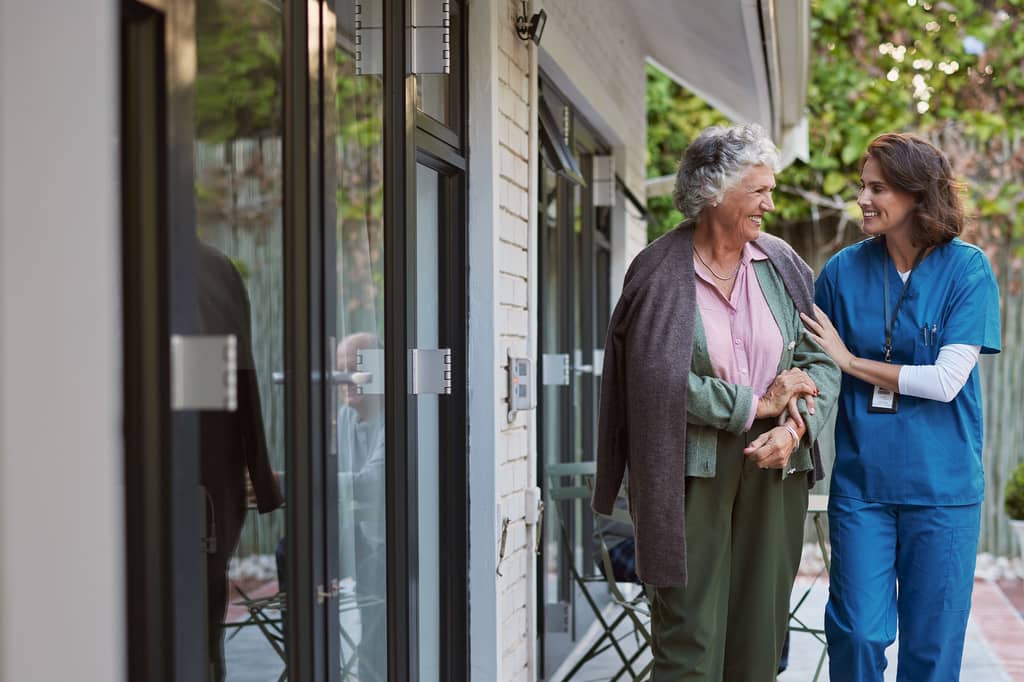 Nurse in blue scrubs supports an older woman as they walk outside together