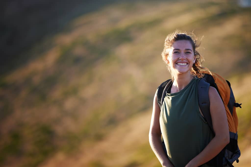 Smiling woman with a backpack hiking on a sunlit grassy hillside