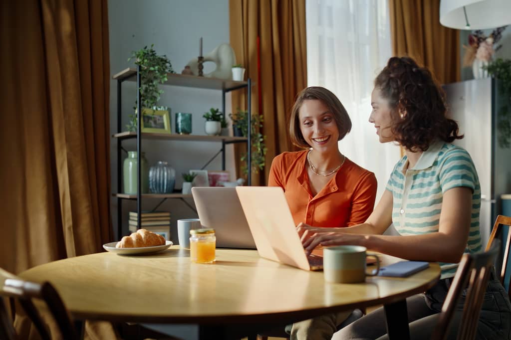 Two women smiling at a laptop at a dining table while comparing options