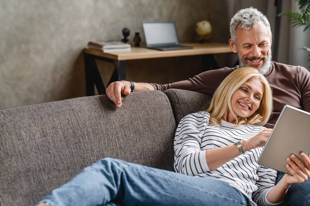 Relaxed older couple smile at a tablet while sitting together on a sofa at home