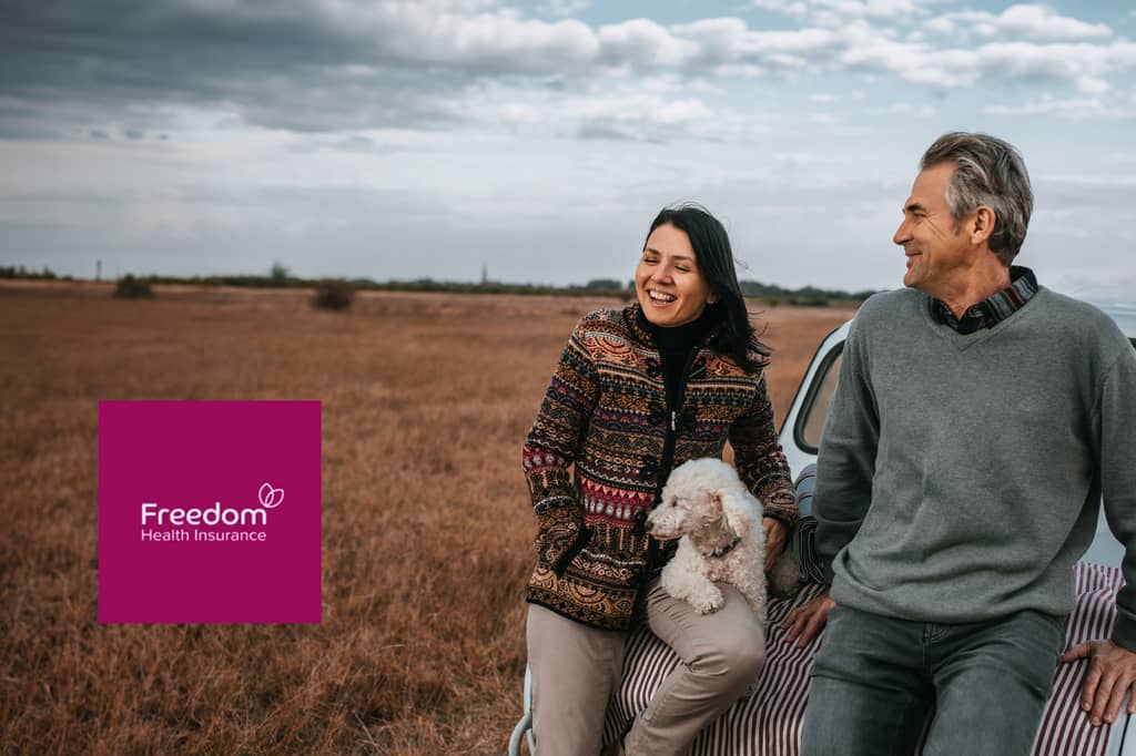 Couple and their dog by a car in a field, with the Freedom Health Insurance logo