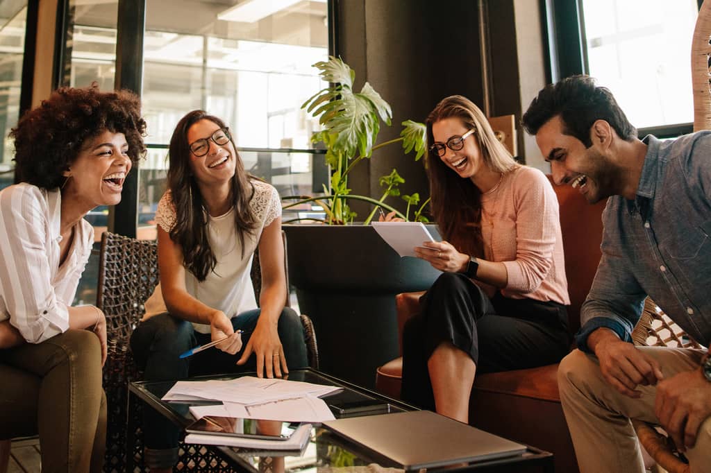 Four young professionals laughing together over paperwork in a relaxed office lounge
