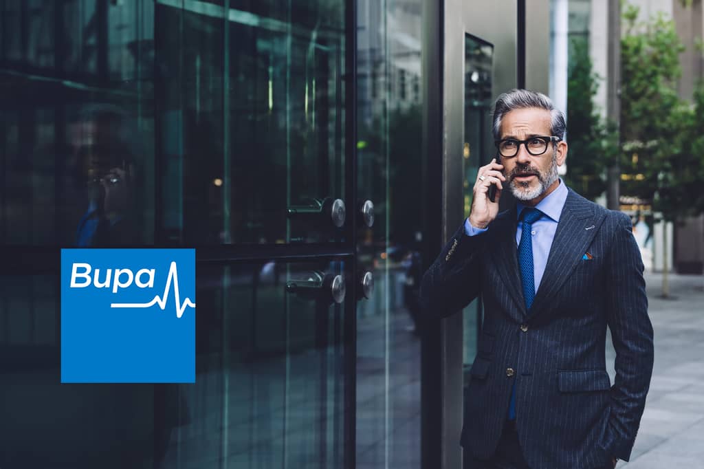 Businessman in a pinstripe suit talks on his phone outside an office building, with the Bupa logo overlaid