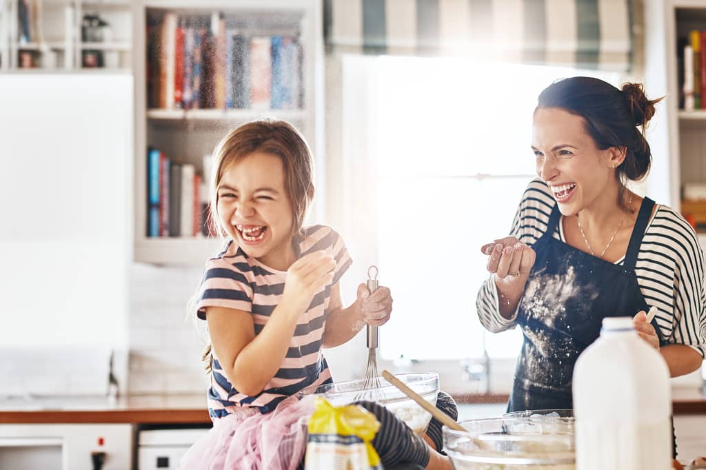 Mother and young daughter laugh while baking together in a bright kitchen