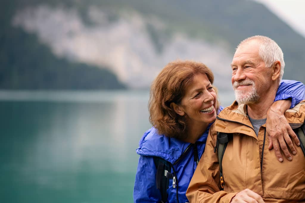 Smiling retired couple in outdoor jackets embracing beside a calm mountain lake