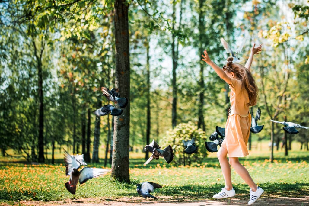 Young girl in a peach dress playing and scattering pigeons in a sunny park