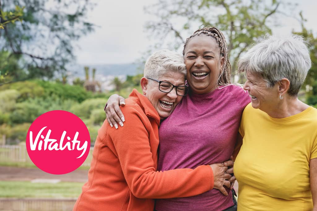Three older women laugh and embrace outdoors, with the Vitality logo overlaid
