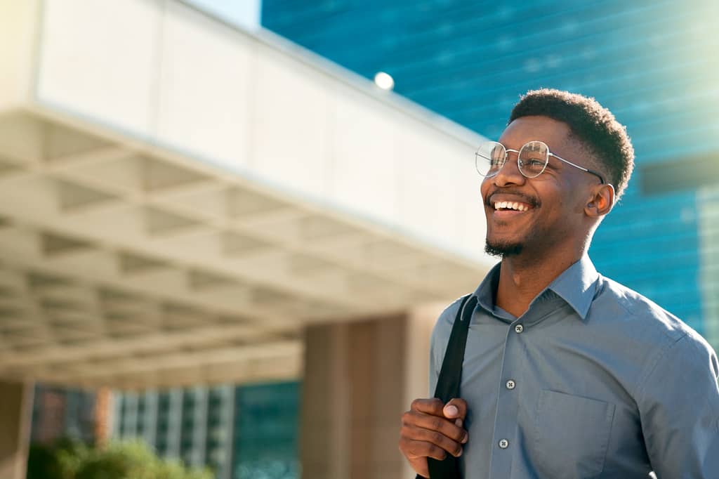 Smiling man with glasses and a shoulder bag stands outside a modern office building