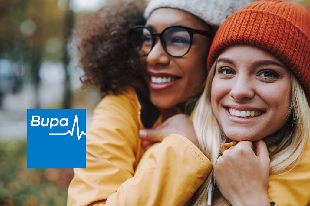 Two friends in colourful coats and hats smile outdoors in autumn, with the Bupa logo overlaid