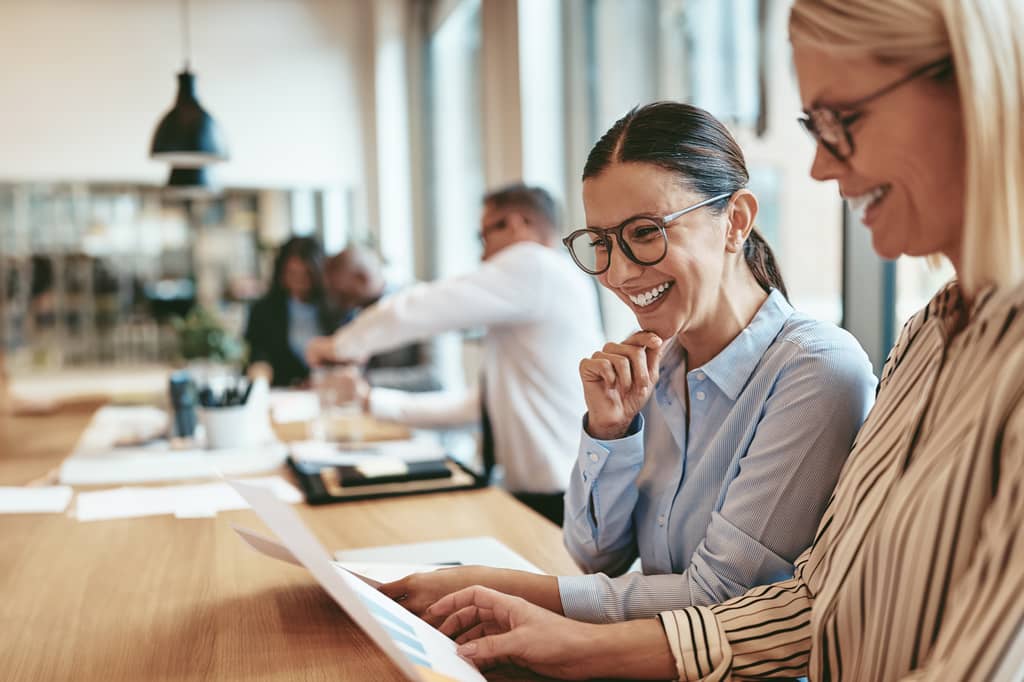 Two colleagues laughing over paperwork at a shared desk in a busy office