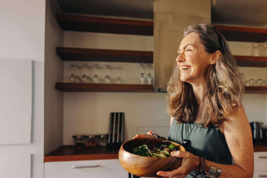 Smiling woman holds a wooden bowl of salad in a sunlit kitchen