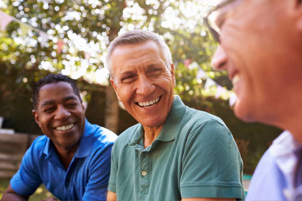 Three middle-aged men laugh together at an outdoor garden gathering