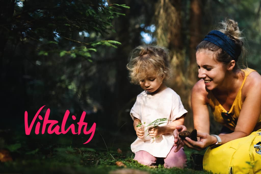 Woman and toddler examine a pine cone and fern together on forest grass. Vitality logo overlaid.