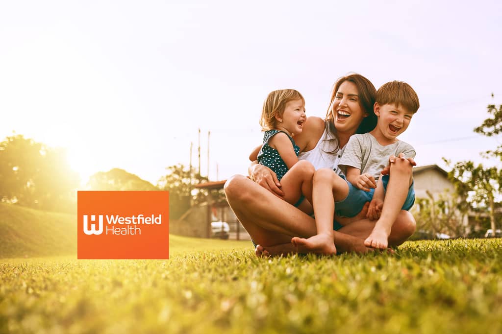 A laughing mother sits cross-legged on grass at sunset, holding her two young children on her lap, all three laughing together, next to the Westfield Health logo.