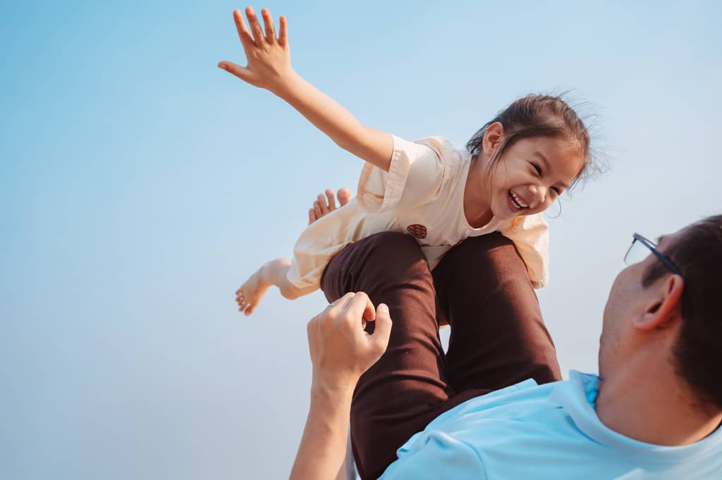 Father lifts laughing young girl overhead, arm outstretched, against a clear blue sky.